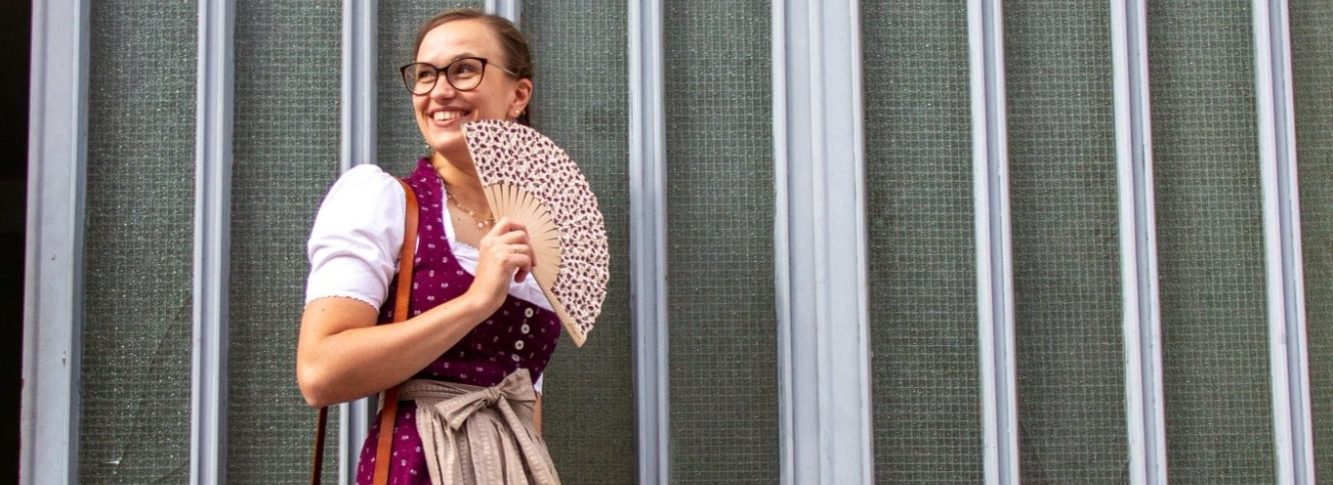 Startbild Aufnahme einer Person mit Trachtenfächer und Bordeaux-farbenem Dirndl vor einer Glaswand